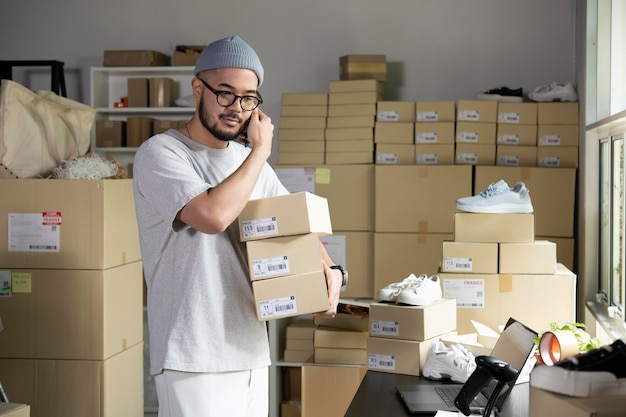 Homme asiatique vendeur en ligne confirmant les commandes du client sur le téléphone et l'ordinateur portable Propriétaire d'entreprise de commerce électronique utilisant le téléphone et détenant des colis dans l'entrepôt du magasin