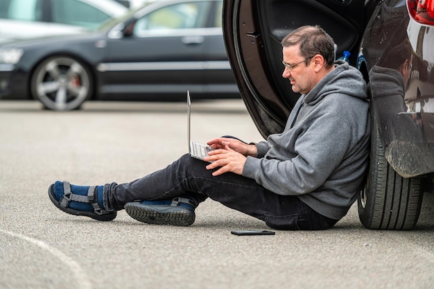 Homme assis sur l'asphalte en voiture et travaillant sur un ordinateur portable travaillant à distance en voyageant