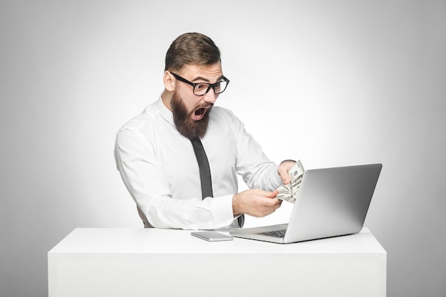 Homme assis au bureau tenant de l'argent avec une bouche surprise et ouverte à la recherche de l'ordinateur portable