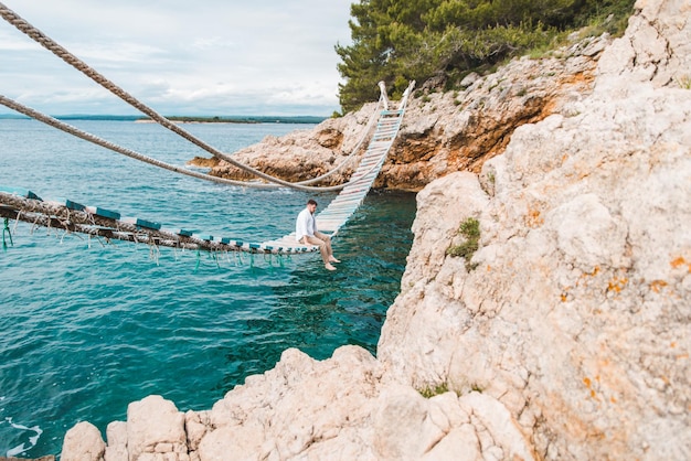 Homme assis au pont suspendu profitant de la vue sur la mer et du calme de la nature l'heure d'été