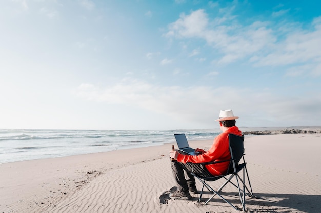 Homme assis sur une chaise de camping sur la plage buvant de la bière avec un ordinateur portable seul profitant