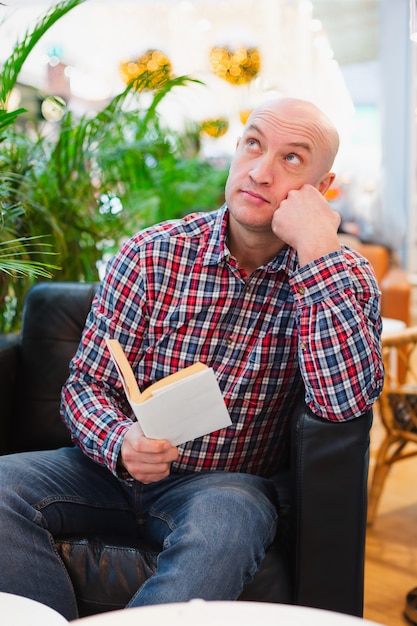 Homme assis sur une chaise dans un appartement lumineux pensif avec un livre dans ses mains