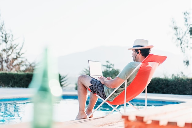 Homme assis sur une chaise de plage au bord de la piscine avec un ordinateur portable seul
