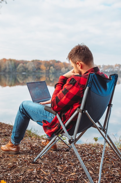 Photo homme assis sur une chaise près du lac d'automne travaillant sur un concept de camping indépendant pour ordinateur portable