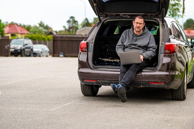 Un homme assis dans le coffre ouvert d'une voiture et travaillant avec un ordinateur portable travail à distance de la technologie mobile