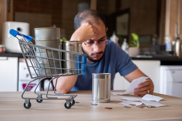 un homme assis dans la cuisine tenant un reçu se sentant frustré en raison de l'inflation et de l'augmentation des besoins quotidiens fournitures pour la maison avec un chariot d'achat devant lui tenant de la nourriture