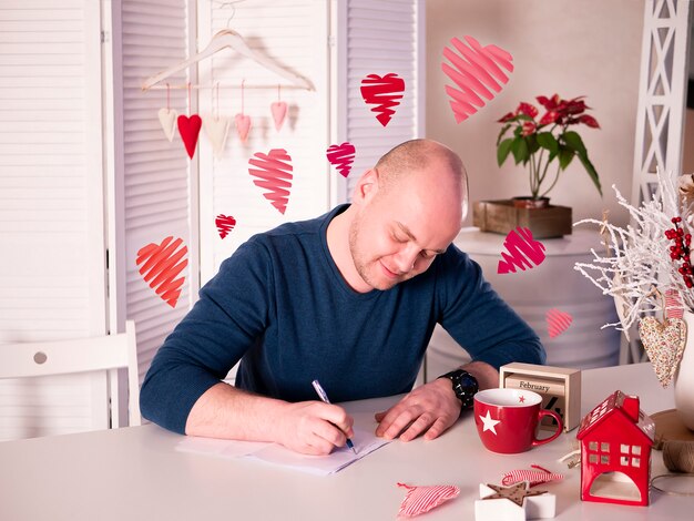 Homme assis et écrivant une lettre d&#39;amour à son amour sur un bel intérieur lumineux avec des cœurs volant autour de lui.