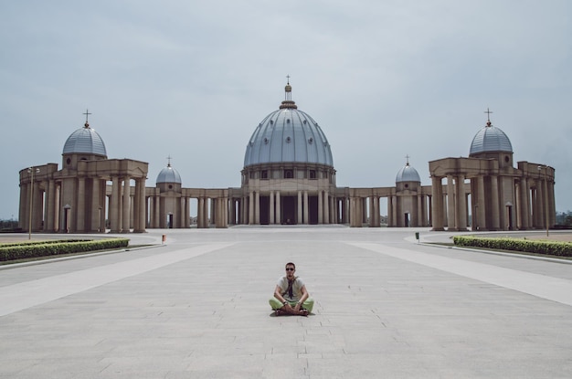 Photo homme assis à l'extérieur de la basilique de notre-dame de la paix cathédrale contre le ciel yamoussoukro afrique