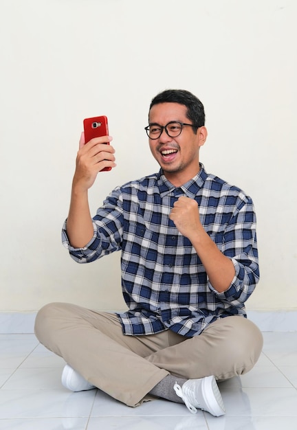 Photo un homme assis les jambes croisées sur le sol montre de l'excitation en regardant son téléphone portable