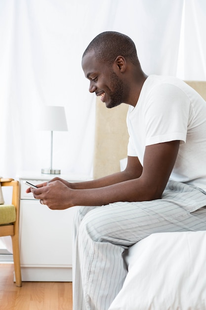 Homme assis sur un lit et SMS sur téléphone portable à la maison