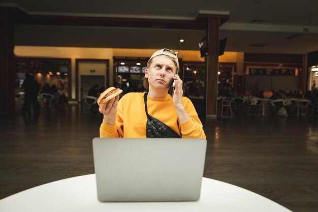 homme assis avec un ordinateur portable dans un café confortable tenant un hamburger à la main et parlant au téléphone