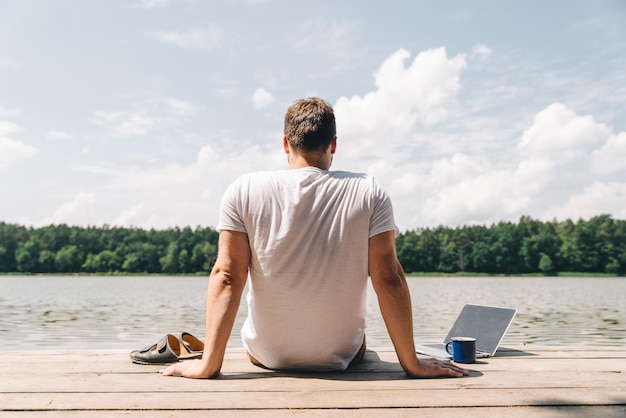 Homme assis avec un ordinateur portable et regardant sur le lac travaillant au concept d'heure d'été de vacances