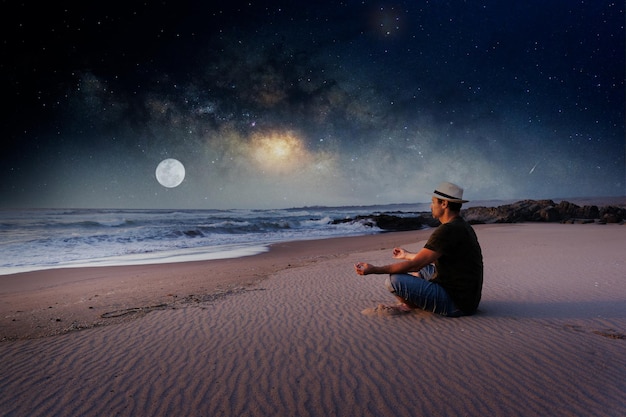 Homme assis sur la plage en position du lotus méditant avec voie lactée et fond de lune