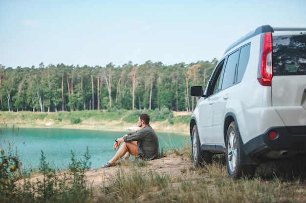 Homme assis près d'une voiture suv blanche au bord regardant le lac avec de l'eau bleue