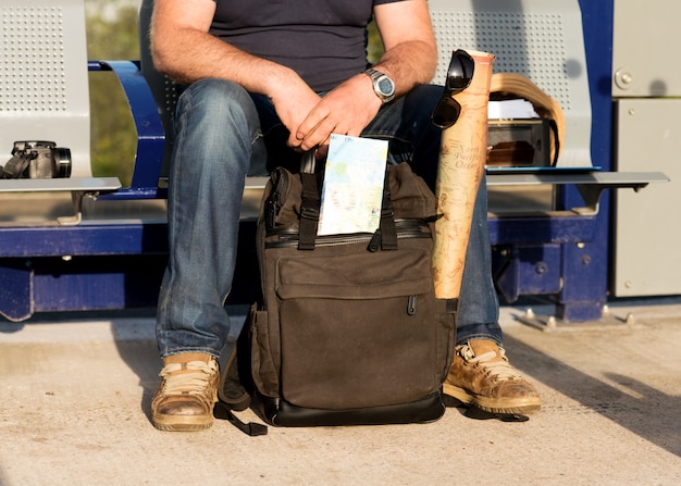 Homme assis avec sac à dos de voyage vert, cartes, lunettes dans une gare vide