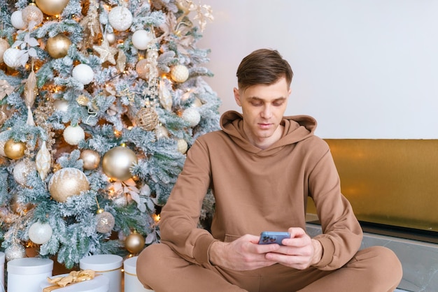 Un homme assis sur le sol près de l'arbre de Noël commande des produits en ligne le vendredi noir