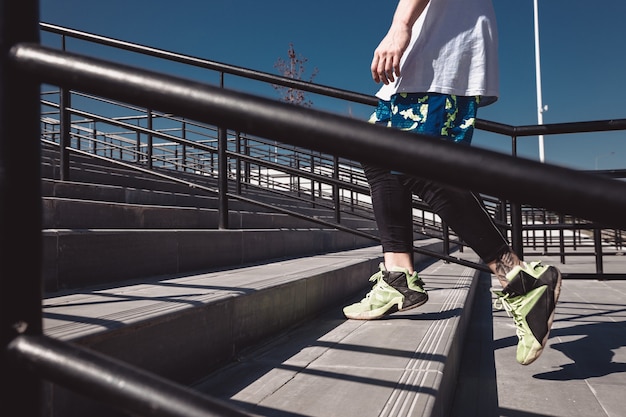 Un homme athlétique avec un bandeau sur la tête vêtu d'un t-shirt blanc, de leggings noirs et d'un short bleu monte les escaliers avec des balustrades noires à l'extérieur par une journée ensoleillée.