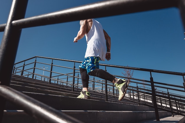 Un homme athlétique avec un bandeau sur la tête vêtu d'un t-shirt blanc, de leggings noirs et d'un short bleu monte les escaliers à l'extérieur par une journée ensoleillée.