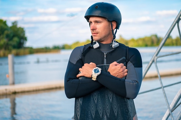 Homme athlétique en combinaison et casque posant avec wakeboard sur la jetée en détournant les yeux