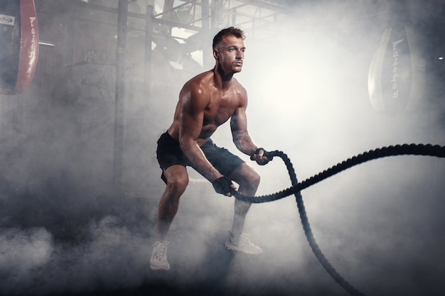 homme athlétique faisant des exercices de crossfit avec une corde dans une salle de sport enfumée