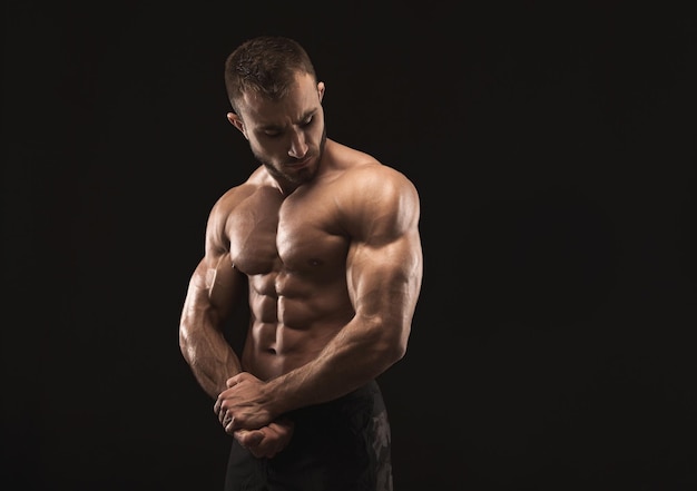 Homme athlétique fort avec torse nu, montrant son corps musclé et ses biceps sur fond noir, discret. Concept de musculation