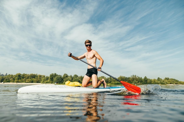 Homme athlétique musclé à lunettes de soleil ramant activement avec une rame assis sur une planche de sup nage sur un étang Plaisirs d'été sur l'eau