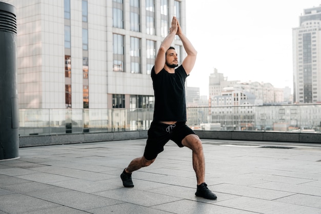 L'homme athlétique pratique la pose Low Lunge avec les bras levés. Jeune homme en t-shirt de sport noir et leggins s'étirant dans une pose de yoga. Concept de sport urbain. Matin échauffement.