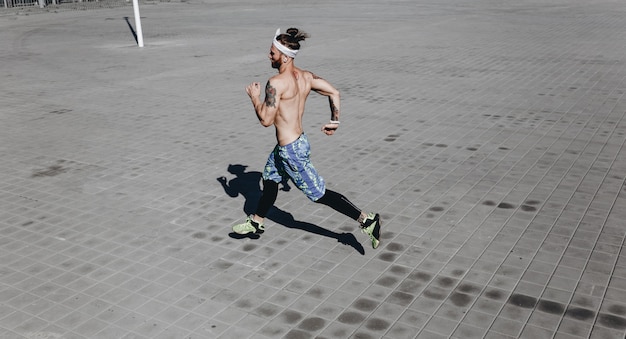 Un homme athlétique avec un torse nu avec des tatouages et un bandeau sur la tête vêtu de leggings noirs et d'un short bleu court sur des dalles par une chaude journée ensoleillée.