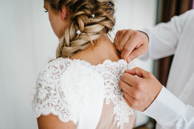 Un homme attache des boutons sur le corset sur la robe. Mariée en robe de mariée blanche avec dentelle debout dans la chambre. Vue arrière.