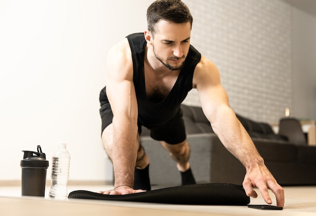 L'homme atteint le téléphone pour changer la musique du téléphone pour le sport. Entraînement sur tapis de yoga à la maison. Exercices du matin avec musique. Activités sportives pendant la quarantaine.