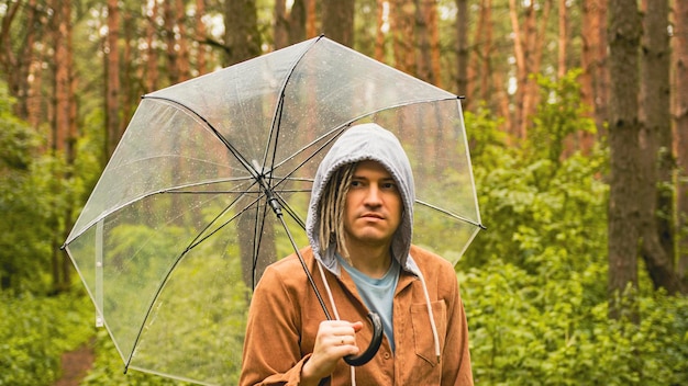 Un homme attentionné avec un parapluie à la main dans les bois Un hipster avec des dreadlocks dans des vêtements décontractés se cache de la pluie sous un parapluie dans les bois