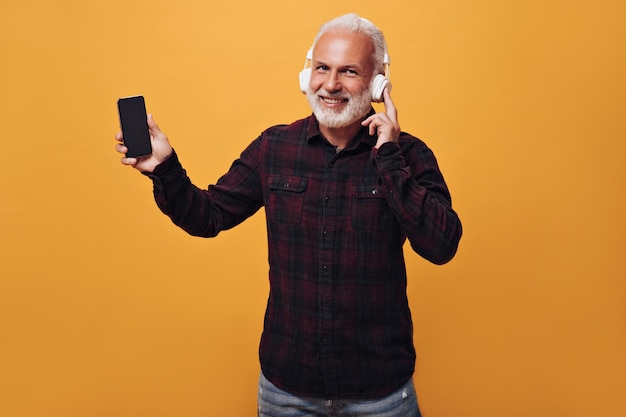 Un homme au casque montre un smartphone sur fond beige Un homme aux cheveux gris pose devant la caméra et sourit