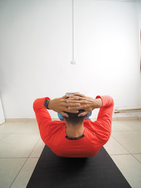 Homme au casque sans fil bleu faisant des exercices de muscles abdominaux allongé sur un tapis de gym. Vue arrière.