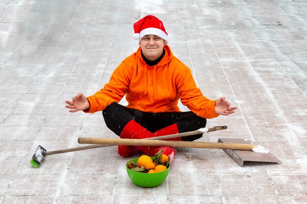 L'homme au chapeau et aux bottes de Noël rouge est assis en position du lotus sur le site de sa cour avec une brosse et une pelle