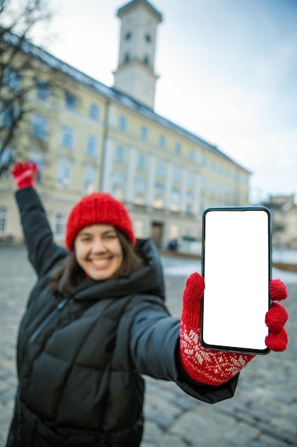Homme au chapeau d'hiver rouge tenant le téléphone avec écran blanc vide vide hôtel de ville européen sur fond