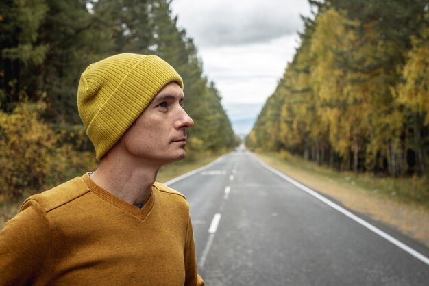 Homme au chapeau jaune et pull sur une route goudronnée vide et lisse le long d'une belle forêt dorée. Concept d'humeur d'automne, de marche, de loisirs de plein air, de voyage et de randonnée