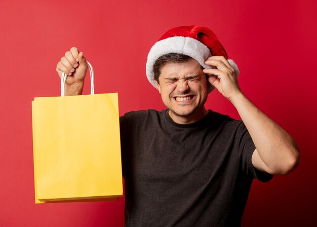 Homme au chapeau de Noël et t-shirt noir avec des sacs à provisions sur l'espace rouge