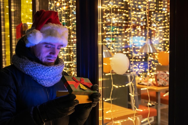 Homme au chapeau de père Noël avec boîte-cadeaux près de la fenêtre du café illuminé