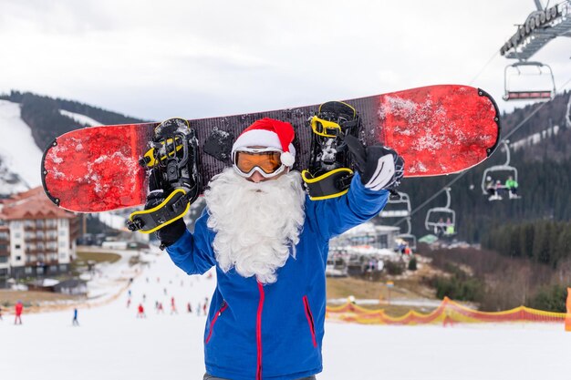 Homme au chapeau de père Noël avec un snowboard dans une station de ski.