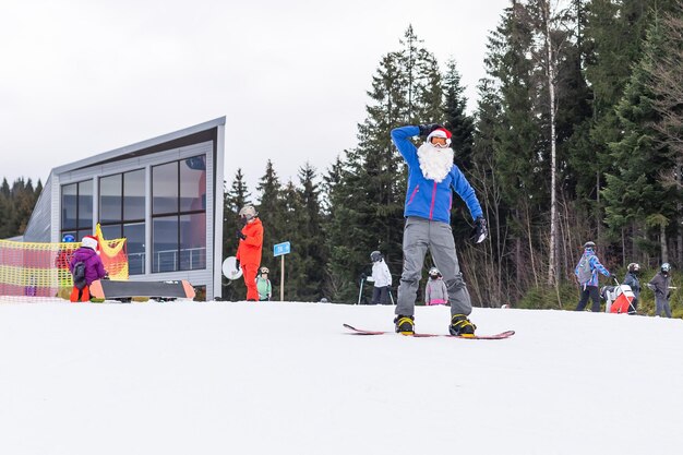 Homme au chapeau de père Noël avec un snowboard dans une station de ski.