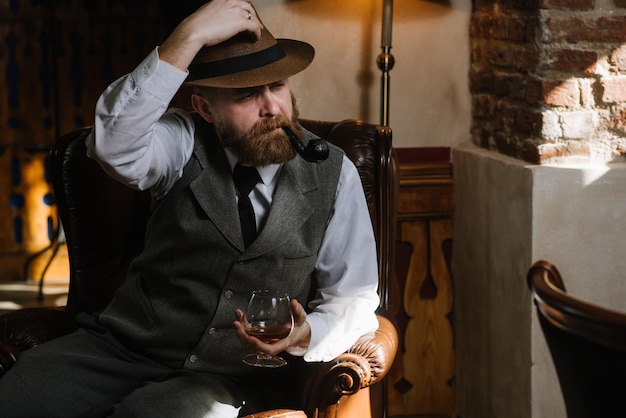 Un homme au chapeau portant un costume vintage tenant une pipe et un verre de whisky assis sur une grande chaise