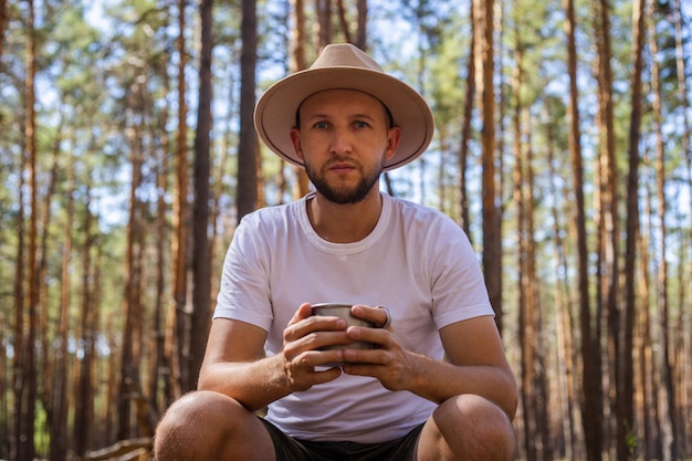 Un homme au chapeau tient une tasse de thé chaud lors d'un voyage de camping Randonnée dans la forêt des montagnes