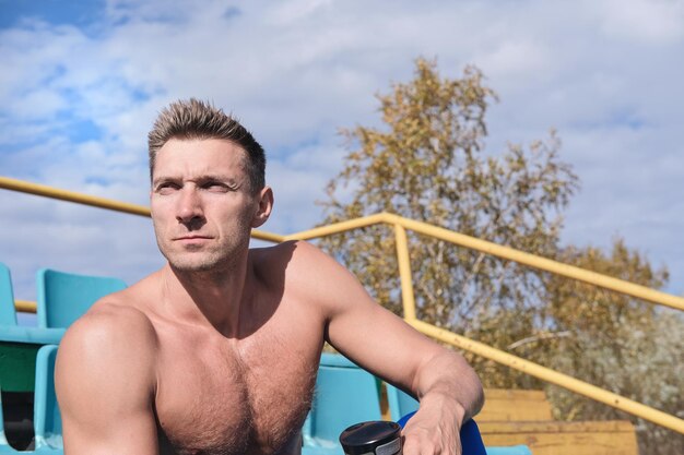 Photo homme au corps tonique d'âge moyen assis torse nu sur la tribune du stade et se reposant après une séance d'entraînement en plein air sport et mode de vie actif bel homme caucasien regardant de côté et se relaxant sur une lumière du soleil