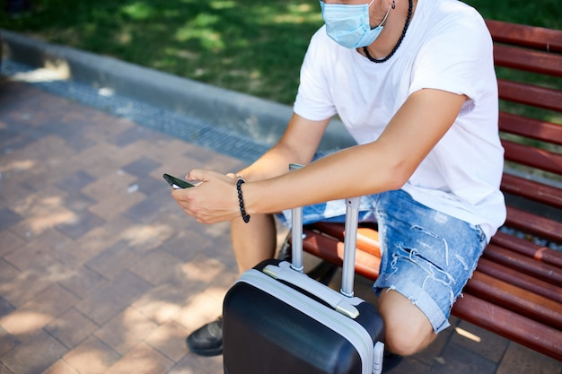 Homme au masque de protection, assis dans le parc en plein air avec une valise et un téléphone portable