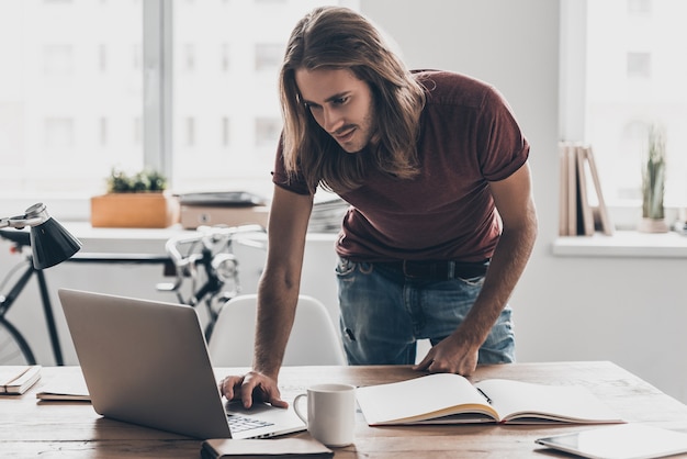 Homme au travail. Beau jeune homme aux cheveux longs regardant son ordinateur portable tout en se penchant au bureau dans un bureau créatif