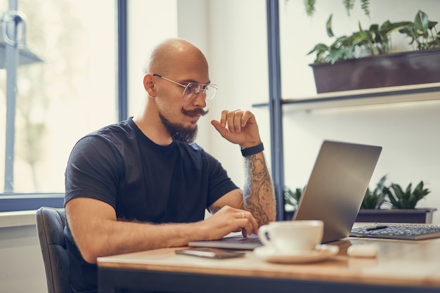 L'homme audacieux avec la moustache et la barbe travaille sur l'ordinateur au bureau à domicile écrivain programmeur freeelancer