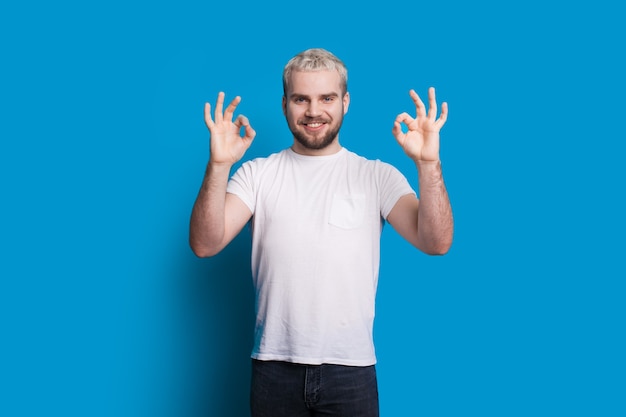 Homme aux cheveux blonds et belle barbe fait signe le signe d'approbation tout en posant dans un t-shirt blanc sur un mur bleu