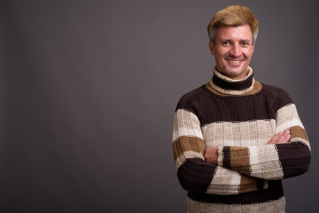 Homme aux cheveux blonds portant un pull à col roulé contre un mur gris