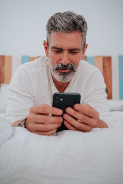 Homme aux cheveux gris dans la cinquantaine allongé sur son lit en regardant le téléphone en se levant