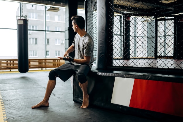 Un homme aux cheveux noirs vêtu d'un t-shirt gris et d'un short noir est assis au bord du ring de boxe et enroule un bandage sur sa main dans la salle de sport.